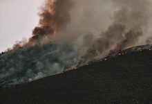 Incêndio florestal se espalha por uma encosta montanhosa, lançando uma espessa coluna de fumaça no céu.