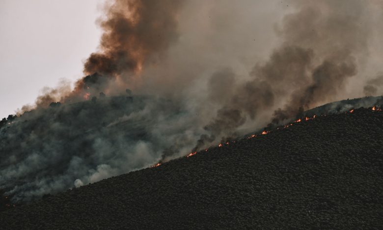 Incêndio florestal se espalha por uma encosta montanhosa, lançando uma espessa coluna de fumaça no céu.